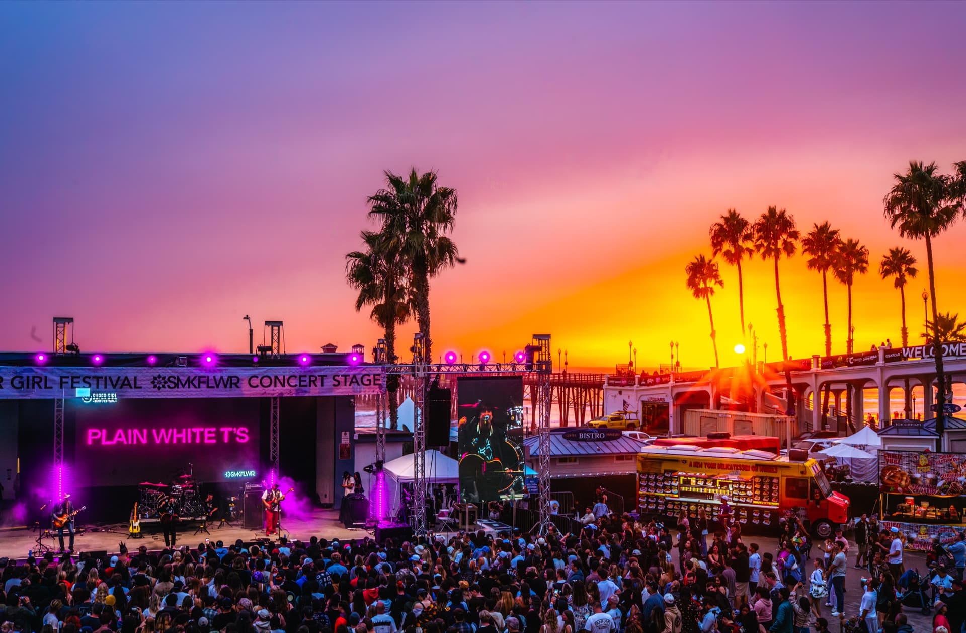 Supergirl outdoor stage at sunset with palm silhouettes, colorful lighting, and a crowd.