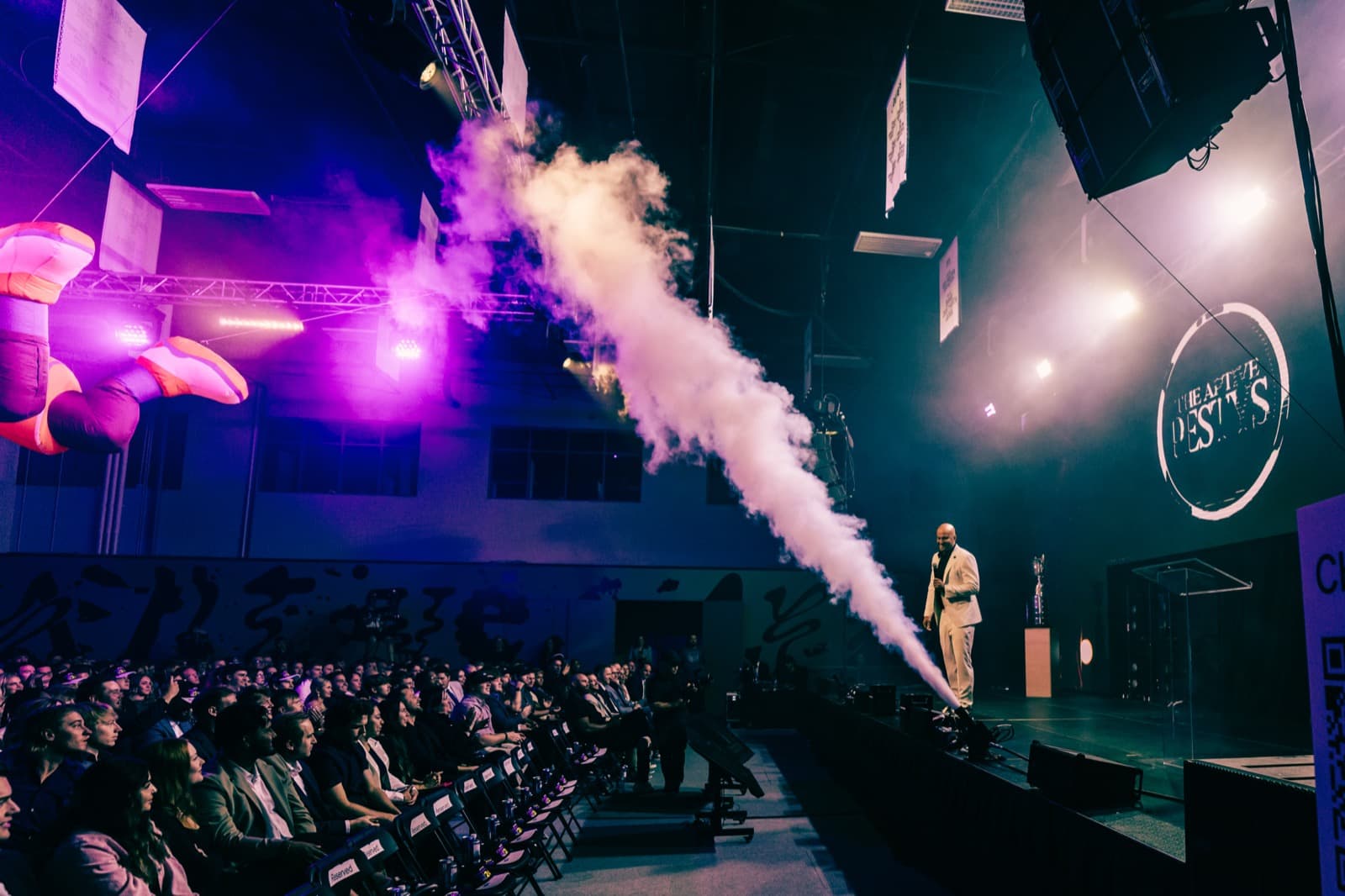 Aptive event stage with fog effects, overhead lighting, and a presenter framed by a large screen.