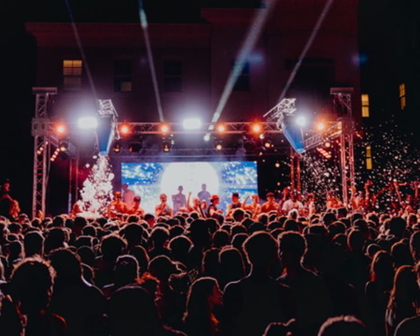 Dense party crowd facing a concert-style stage with white beams and haze.